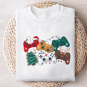 May include: White sweatshirt featuring a festive design of colorful game controllers. The controllers are decorated with Christmas elements like a Santa hat, snowflakes, and reindeer antlers. A Christmas tree and string lights are also included.