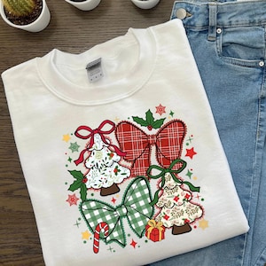 Pode incluir: Camisola branca com um design temático de Natal. O design inclui duas árvores de Natal com padrões florais e dois grandes laços em xadrez vermelho e verde. Uma bengala de doces e uma caixa de presente também estão incluídas. A camisola é combinada com jeans azuis.