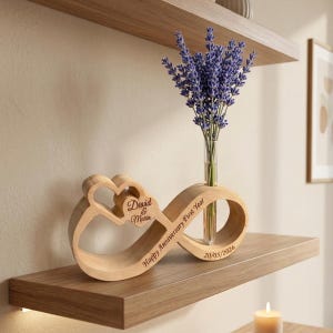 Puede incluir: Un jarrón de madera con forma de infinito con un tubo de vidrio transparente que contiene lavanda morada. El jarrón tiene texto grabado que dice "David & Maria" y "Happy Anniversary First Year 20/05/2026". El jarrón está sobre una estantería de madera.