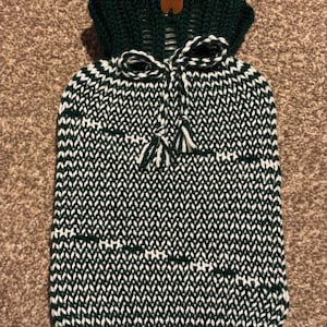 Puede incluir: Una funda de botella de agua caliente tejida en verde oscuro y blanco. La funda presenta un patrón texturizado, un lazo y una borla. La parte superior tiene una etiqueta que dice "Happy Cat".