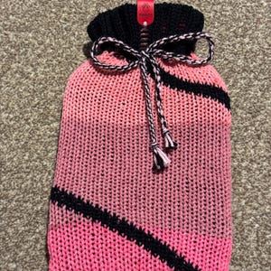 Puede incluir: Funda de botella de agua caliente de punto rosa y negro. La funda tiene una parte superior de punto negro y una franja diagonal negra en la parte delantera. Un cordón negro y blanco está atado con un lazo en la parte superior. Una etiqueta roja está adjunta.