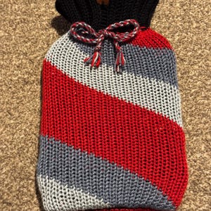 May include: A knitted hot water bottle cover with a black top and a diagonal pattern of red, grey, and white stripes. A decorative bow with red, white, and grey strings is tied at the top. A small brown tag with the text "Happy Cat" is attached.