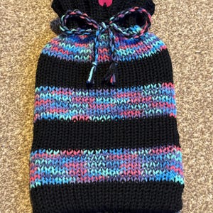 May include: A black knitted hot water bottle cover with horizontal stripes in shades of blue, pink, and purple. The top is gathered and tied with a matching cord. A pink tag with the text "Happy Cat" is attached.