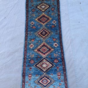 May include: A rectangular runner rug featuring a vibrant blue field with a central vertical row of six interconnected diamond-shaped medallions. The rug displays intricate geometric patterns, stylized animal motifs, and a dark red border.