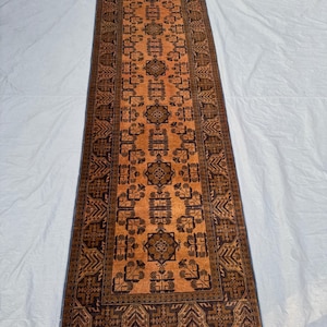 May include: A long, rectangular rug with an intricate geometric pattern. The rug features a warm orange-brown central field with dark brown and black detailing. The edges are bordered with a complex design, and the ends have white fringe.