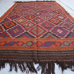 Puede incluir: Una alfombra rectangular tejida a mano con un patrón geométrico. La alfombra presenta un diseño de diamantes repetidos en tonos de rojo, naranja, verde y morado. El borde es marrón oscuro con flecos en el borde inferior. Un textil decorativo.