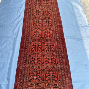 Puede incluir: Una alfombra larga y rectangular con un detallado patrón geométrico en tonos rojos, negros y marrones. La alfombra presenta un diseño central con motivos repetidos y un borde con flecos. La alfombra está colocada sobre una superficie azul.