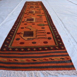 May include: An orange runner rug with a camel design. The rug has a dark brown border with geometric patterns and a fringe at the end. The camel figures are in a lighter brown color.