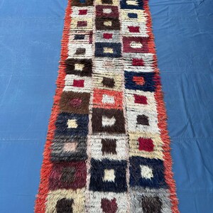 Puede incluir: Una alfombra rectangular hecha a mano con un patrón repetido de cuadrados en varios colores. Los cuadrados incluyen tonos de rojo, marrón, beige y azul. La alfombra tiene una textura peluda y un borde rojo. Esta alfombra es adecuada para un pasillo o entrada.