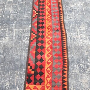 Puede incluir: Una alfombra larga y rectangular con un patrón geométrico. La alfombra presenta un diseño central de formas rojas, negras, naranjas y grises. El borde es de color marrón oscuro. La alfombra probablemente está hecha de material tejido.