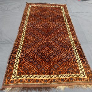 Puede incluir: Alfombra rectangular con un patrón geométrico en tonos marrón, naranja y azul. La alfombra presenta un diseño central de diamantes y un borde crema con un patrón repetitivo. Se ven flecos en ambos extremos.