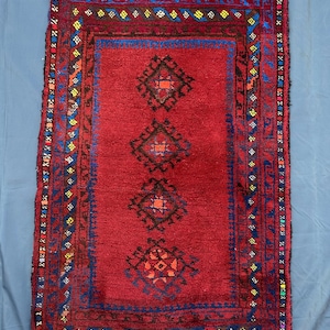 Puede incluir: Una alfombra rectangular, tejida a mano, con un campo rojo intenso y patrones geométricos intrincados. La alfombra presenta un diseño central de tres formas de diamante en negro y naranja, enmarcado por un borde azul con acentos coloridos. Los bordes tienen un acabado con flecos.