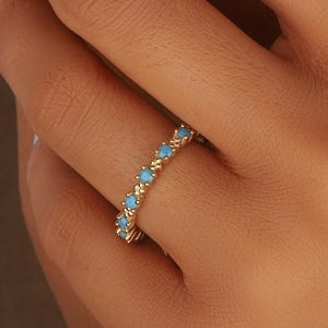 Op de afbeelding: Een gouden ring met kleine turkooizen stenen in een herhalend patroon. De ring zit om een vinger, met de hand en andere vingers zichtbaar. De nagels zijn wit gelakt.