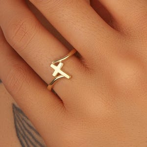 May include: A gold-colored ring with a cross design. The ring is worn on a finger. The cross is the focal point of the ring, and the band is thin and delicate. The ring is a piece of jewelry.