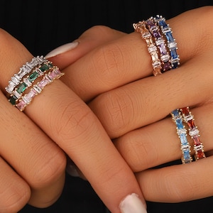 May include: Close-up of several rings adorned with colourful rectangular gemstones. The rings display emerald green, pink, blue, red, and purple stones. The rings are worn on fingers, highlighting the jewellery's design and colour variations.