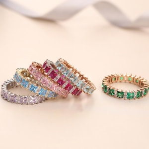 Op de afbeelding: Een verzameling kleurrijke eternity-ringen. De ringen hebben verschillende gekleurde edelstenen, waaronder paars, blauw, roze, rood en groen. De ringen zijn gezet in goudkleurige banden en worden gepresenteerd op een lichtgekleurd oppervlak.