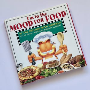 May include: A cookbook titled "I'm in the MOOD FOR FOOD" featuring Garfield the cat in a chef's hat. The cover displays Garfield holding a spatula and wooden spoon, surrounded by various food items like pizza, salad, and pancakes.