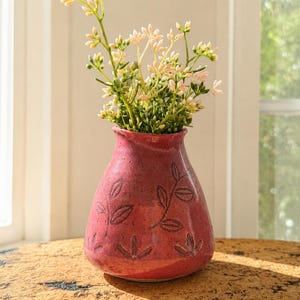 May include: A small, rounded ceramic vase with a glossy, speckled raspberry-red glaze. The surface features hand-carved, dark-outlined leaf and floral motifs. A cluster of delicate, light-coloured artificial flowers with green stems sits inside.
