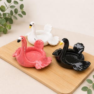 May include: Three ceramic bird-shaped trinket dishes in pink, white, and black. Each dish features a sculpted bird with a long neck, a small yellow beak, and spread wings forming the bowl. The pink bird has white dots, the white bird has black dots, and the black bird has a solid finish. These decorative dishes are arranged together.