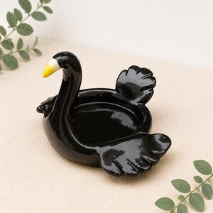 Peut inclure: Un vide-poche en céramique noire brillante en forme de cygne avec une finition lisse. Le cygne présente un bec jaune vif avec une pointe blanche, un long cou et des ailes stylisées qui forment le bassin circulaire pour les petits objets.