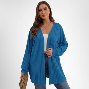 May include: A long-sleeved, open-front cardigan in a vibrant blue hue. The cardigan features a ribbed texture and a relaxed fit. The model is wearing the cardigan over a white top and blue jeans. A tan clutch is held in the left hand.
