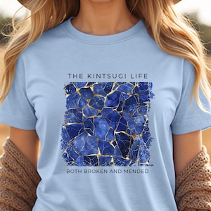 May include: Light blue t-shirt featuring the text "THE KINTSUGI LIFE" above a square design of blue mosaic pieces with gold accents. The text "BOTH BROKEN AND MENDED" is below the design.