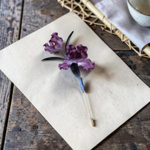 May include: A lapel pin featuring two deep purple orchid-like flowers with ruffled petals and a small yellow centre. The floral element is attached to a thin, straight gold-toned metal pin stem with a pointed end and a small metal stopper.