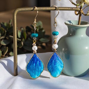 May include: Dangle earrings with gold-tone hooks and accents. Each earring has a teardrop-shaped blue and gold pendant, a white pearl, and a turquoise bead. The earrings are displayed on a gold-coloured metal stand.