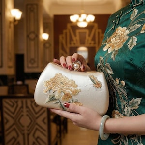 May include: Ivory satin clutch bag with gold floral and butterfly embroidery. The bag has a pearl clasp. The person holding the bag wears a green dress with gold floral embroidery and a jade bracelet.