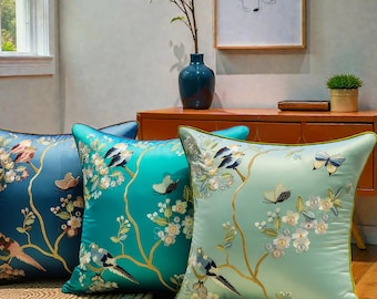 May include: Three decorative throw pillows in shades of teal and blue. Each pillow features embroidered birds, butterflies, and floral branches. The pillows have a satin-like sheen and are arranged on a neutral-toned rug.