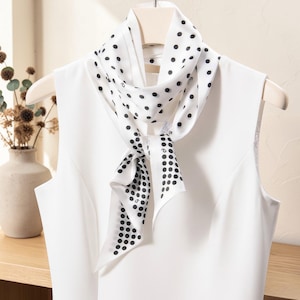 May include: A white silk scarf with a black polka dot pattern is draped over a white sleeveless top. The scarf is tied in a knot, and the ends hang down. The scarf is hanging on a wooden hanger.
