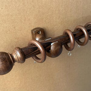 May include: A wooden curtain rod set with a dark brown finish. The set includes a rod, decorative finials, rings, and mounting hardware. The rod is approximately 152 cm long.