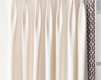 May include: Cream-colored pleated curtains hanging from a gold-toned rod. The curtains have a tailored look with vertical folds. A decorative panel with a geometric pattern in a darker shade is visible on the right side.
