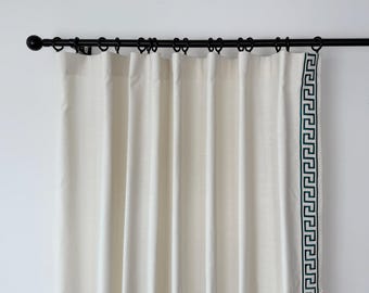 May include: Off-white curtain panel with a dark green Greek key border, hanging from a black metal rod with rings. The curtain is made of a textured fabric and is partially drawn, showing vertical folds.