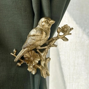 May include: A gold-coloured metal curtain tieback depicting a bird perched on a flowering branch. The bird is detailed with textured feathers and sits on a branch with two flowers and several leaves. The tieback is attached to a dark green curtain.