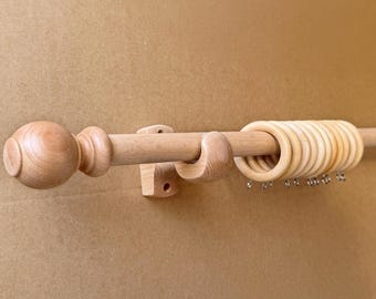 May include: A light-colored wooden curtain rod with a decorative finial and rings. The rod is mounted on a wall with wooden brackets. The rings have small metal hooks for hanging curtains. The finial is a rounded shape.