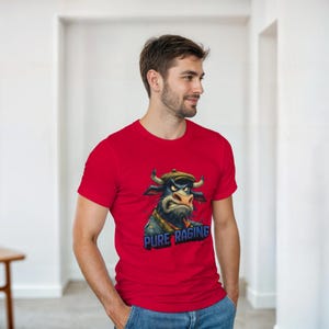 Op de afbeelding: Rood t-shirt met een cartoon stier met een pet en sjaal, met de tekst "PURE RAGING". De stier heeft een boze uitdrukking. Het t-shirt is gemaakt van een zacht materiaal.
