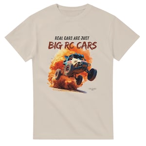 Peut inclure: T-shirt beige avec le texte "REAL CARS ARE JUST BIG RC CARS" au-dessus d'une illustration d'une voiture RC bleue et blanche. La voiture RC a des roues rouges et est en l'air, soulevant de la poussière.