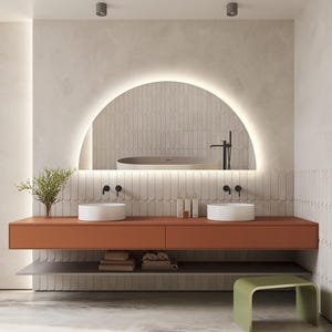 May include: Modern bathroom design featuring a large, illuminated arched mirror above a burnt orange floating vanity with two white vessel sinks. The space includes a gray shelf, a green stool, and a neutral tile backsplash. A bathtub is visible in the mirror.