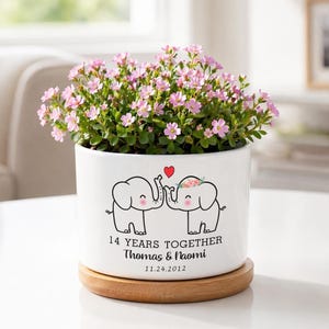 May include: A white cylindrical ceramic planter on a round wooden base, containing a small plant with pink flowers. The planter features a graphic of two elephants with a red heart, and the text 14 YEARS TOGETHER, Thomas & Naomi, 11.24.2012.