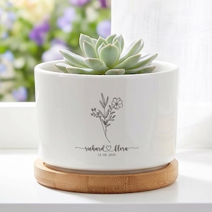 Peut inclure: Un pot de fleurs cylindrique en céramique blanche contenant une plante succulente vert clair. Le pot présente un dessin au trait noir de fleurs sauvages, les noms "richard" et "flora" séparés par un cœur, et la date "12.08.2021". Il repose sur une base ronde en bambou.