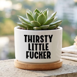 May include: A white ceramic cylindrical planter with a bamboo saucer base, featuring the black text THIRSTY LITTLE FUCKER printed on the front. A green succulent plant is potted inside the container, which has a smooth, glossy finish.