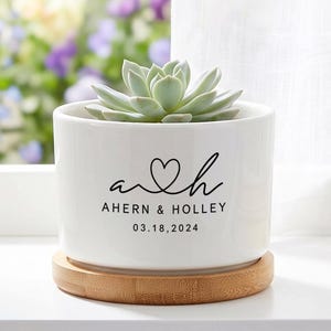 May include: A white ceramic cylindrical planter with a light green succulent plant. The planter features black text reading a heart h, AHERN & HOLLEY, and 03.18.2024. It sits on a circular light brown wooden base with a smooth, natural grain finish.