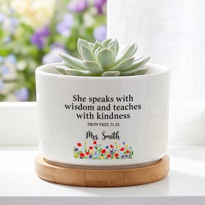 May include: A white ceramic cylindrical planter with a light green succulent plant. The planter features the black text 'She speaks with wisdom and teaches with kindness PROV FREE 31.26 Mrs. Smith' above a colorful floral graphic. It sits on a bamboo base.