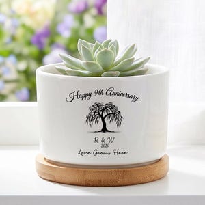 May include: White ceramic plant pot with a succulent. The pot sits on a wooden base and has the text "Happy 9th Anniversary", a tree illustration, and the names "R & W" with the year "2026" and the phrase "Love Grows Here".