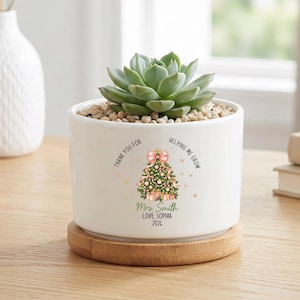 May include: A white ceramic cylindrical planter with a bamboo base containing a green succulent. The front features a printed Christmas tree graphic with a pink bow, gold stars, and the text: Thank you for helping me grow, Mrs. Smith, Love, Sophia, 2024.