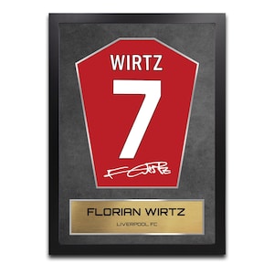 Könnte beinhalten: Gerahmtes Kunstwerk mit einem roten Trikot-Design, das "WIRTZ" und die Nummer "7" in Weiß zeigt. Eine goldfarbene Plakette darunter trägt die Aufschrift "FLORIAN WIRTZ" und "LIVERPOOL FC". Der schwarze Rahmen präsentiert ein Fußball-Thema.