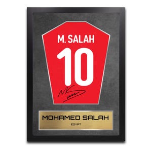 May include: Framed artwork featuring a red soccer jersey design with the name "M. SALAH" and the number "10" in white. A gold-colored plaque at the bottom reads "MOHAMED SALAH" and "EGYPT". The artwork is encased in a black frame.