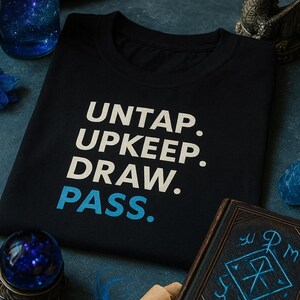 May include: Black t-shirt with the words "UNTAP. UPKEEP. DRAW. PASS." in white and blue. The shirt is surrounded by fantasy-themed items, including a spell book, scrolls, and a decorative dragon statue.