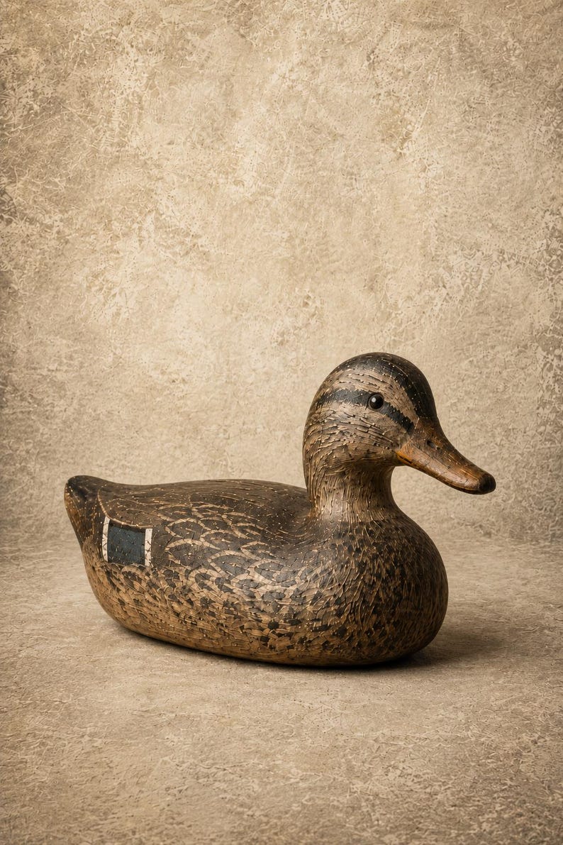 Pode incluir: Um chamariz de pato em madeira esculpida com um padr&atilde;o de plumagem texturizado em castanho e preto, uma cabe&ccedil;a lisa com uma faixa escura no olho e uma mancha retangular azul na asa. A superf&iacute;cie apresenta entalhes detalhados de penas.